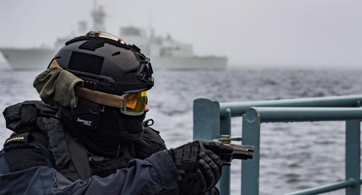 Une personne en tenue tactique tenant une arme de poing sur un bateau avec un navire en arrière-plan.