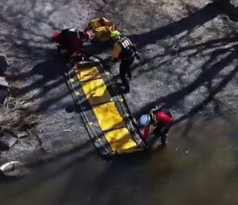 Rescue team with a stretcher beside an overturned car by a rocky terrain.