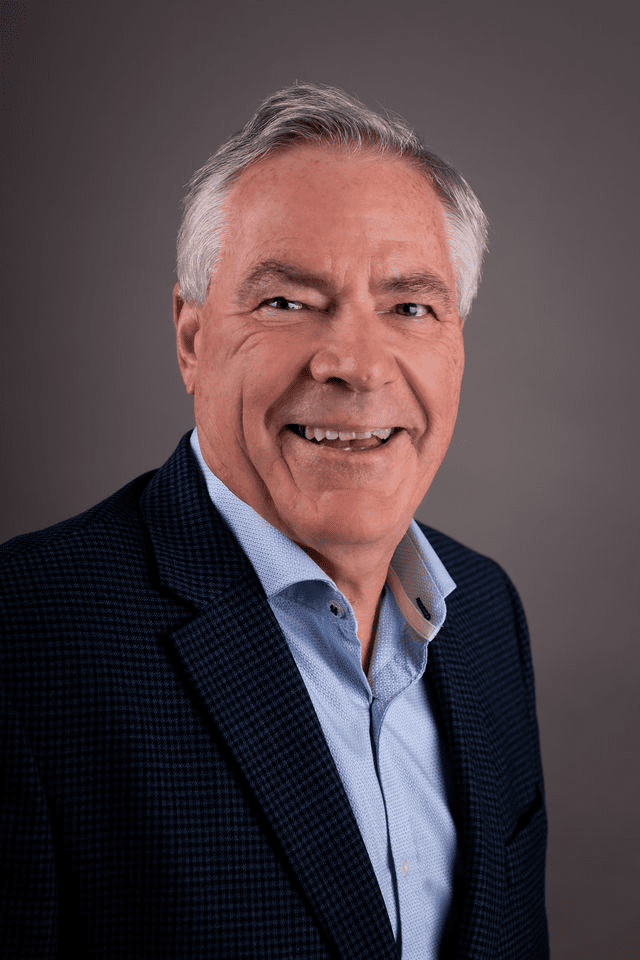 Homme vêtu d'un blazer à motifs bleus et d'une chemise bleu clair, cheveux gris, sur fond sombre.